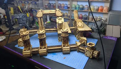 A detailed assembly of the TT Combat Sector 4 Plasma Relay is displayed, featuring intricate wooden structures with multiple towers and connecting pathways. The design includes cylindrical towers with circular tops and various platforms, all set against a blue cutting mat. In the background, jars of paint and tools are visible, indicating a workspace dedicated to tabletop gaming and model building.