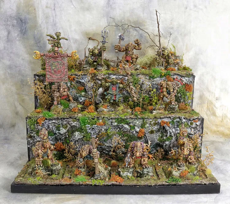 A detailed display features ten hand-sculpted, multi-part miniatures inspired by Celtic mythology, arranged on a tiered rocky terrain adorned with moss and foliage. The miniatures, showcasing intricate designs and characterful features, include various creatures and banners, with some holding weapons and others displaying unique shields. The backdrop consists of a textured surface that enhances the mystical atmosphere, reflecting the coastal marshes and ruins theme of the Famhairean campaign.