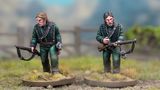 Two detailed miniatures of soldiers from the Sharpe’s Rifles series are displayed on textured bases, set against a blurred natural background. Both figures wear green uniforms with black crossbelts; one holds a rifle and has a serious expression, while the other appears to be in a more contemplative pose. The bases feature tufts of grass, enhancing the realistic terrain effect.