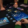 Two people playing a space-themed board game on a table. The game board features a blue space map with planets, ships, and icons. Players use round tokens and cards. One hand holds a rulebook with visible text: "RULEBOOK" and "STAR TREK." Wristbands and scattered game pieces are present.