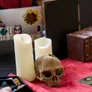 A tabletop scene features two white candles and a decorative skull, set against a red cloth. Nearby, a black tray holds various polyhedral dice, while a small wooden chest is partially visible in the background. The overall arrangement evokes a gothic and mysterious atmosphere, fitting for a vampire-themed gaming event.