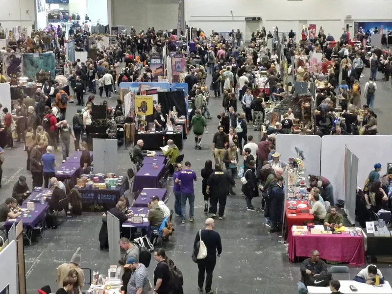 Crowded convention hall with people browsing vendor booths and gaming tables.