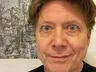 A close-up portrait of a man with light brown hair and striking green eyes, smiling slightly. He is wearing a black shirt and is positioned in front of a detailed, artistic background featuring a complex, fantastical structure, possibly a building or landscape. The overall tone of the image is casual and inviting, with a focus on the subject's expressive features.