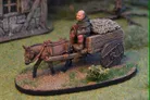 A miniature scene features a small cart being pulled by a brown horse, with a seated figure dressed in rustic clothing, holding the reins. The cart is laden with a bundle of hay or straw, and the base is detailed with grass and small plants. In the background, a stone building adds to the fantasy setting, enhancing the overall atmosphere of a quaint village scene.