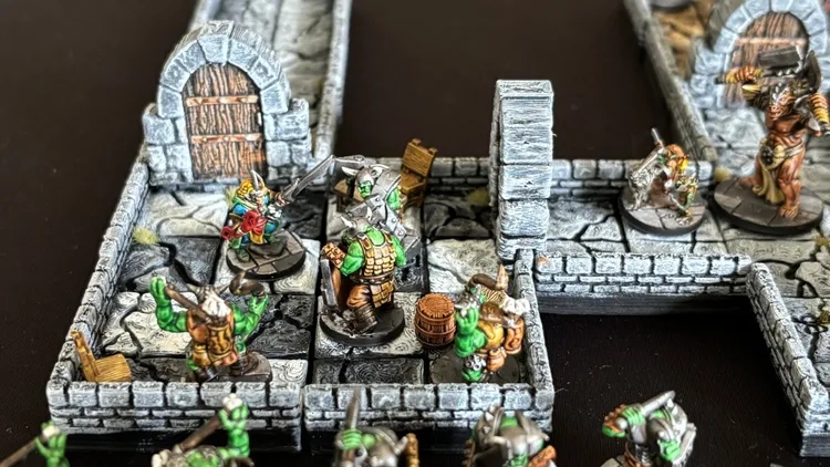 A detailed 15mm scale dungeon scene features a modular layout with stone walls and various tiles. In the foreground, a group of four heroes, including a wizard and a warrior, confront a pair of armored orcs, while scattered barrels and crates add to the environment. The dungeon's intricate design showcases a mix of characters and monsters, enhancing the tabletop gaming experience.