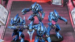 Four Sangheili warriors are depicted in striking blue armor, each wielding different weapons including a needler, plasma rifle, pulse carbine, and energy sword. They are positioned on a futuristic, metallic battlefield with a red and gray color scheme, featuring geometric structures in the background. The warriors exhibit dynamic poses, showcasing their formidable presence as mercenaries in the Halo Flashpoint universe.