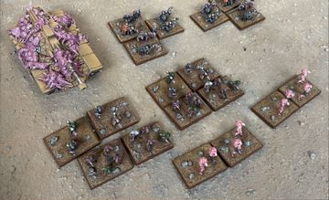 A collection of Zayken miniatures from Void Scar Miniatures is displayed on a textured surface. In the foreground, several bases feature hordes of zombified figures in various colors, including green, purple, and pink, representing the Neophyte Mobs. To the left, a tank is adorned with grotesque, bloated creatures, suggesting a fusion of machine and undead, while additional bases of zombies are scattered throughout the scene, emphasizing the overwhelming numbers of the Zayken forces.