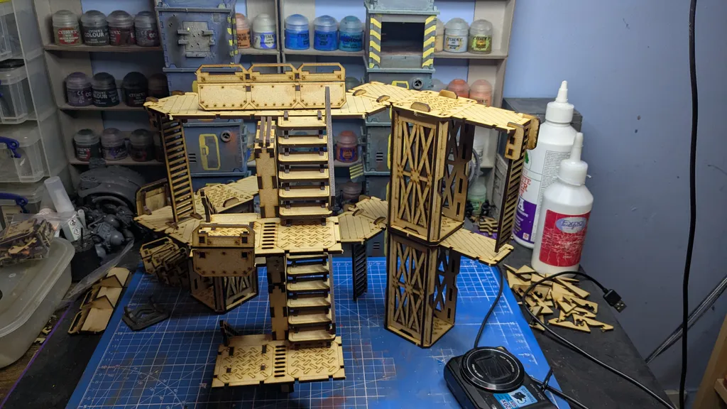 A detailed assembly of modular terrain pieces made from laser-cut wood, featuring multiple levels, staircases, and platforms. The structure includes various textures and patterns, with a mix of open and enclosed areas, showcasing a futuristic industrial design. In the background, paint bottles and tools are visible, indicating a workspace for painting and customizing the terrain.
