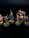 A group of eight intricately painted Moonstone miniatures stands against a black background. The characters include a muscular figure with a weapon, a green-skinned goblin in colorful attire, a knight in armor, and various other fantastical beings, each displaying unique features and vibrant colors. They are positioned on circular bases adorned with tufts of grass and small flowers, showcasing the detailed artistry of the miniatures.