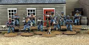 A group of ten detailed 28mm scale miniatures representing dismounted US Cavalry and Buffalo Soldiers stands in front of a wooden building with a red door. The miniatures are posed with various weapons, including rifles and a bugle, showcasing a mix of uniforms and accessories, such as scarves and hats. A small beagle and a cactus are also present among the figures, all mounted on 25mm round bases with a sandy terrain effect.