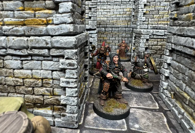 A detailed scene inside a dungeon features four painted miniatures positioned among stone walls and a flagstone floor. In the foreground, a character with long black hair and a fierce expression holds a weapon, flanked by a bearded figure in a red hat and a woman in a brown dress. Another character in armor stands nearby, creating a sense of tension in the dimly lit environment. The walls are textured with a weathered stone appearance, enhancing the dungeon's atmosphere.