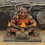 A large, menacing demon figure sits cross-legged, with prominent horns and glowing red eyes, surrounded by flames emanating from two braziers on either side. On its lap, a smaller figure appears to be offering a tribute, while another figure climbs onto the demon's shoulder, creating a dynamic scene. The base features intricate stonework, enhancing the overall fantasy aesthetic.