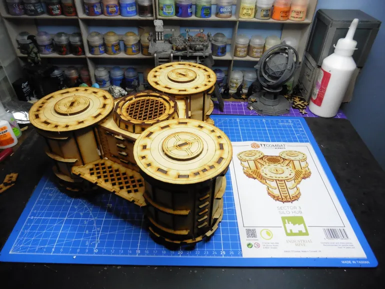 A detailed model of the Sector 3 Silo Hub from TT Combat is prominently displayed on a blue cutting mat. The structure features multiple cylindrical silos with intricate wooden textures and circular tops, showcasing a combination of open latticework and solid panels. In the background, various paint pots and a few other miniature models are visible, along with the instruction sheet for the Silo Hub, which includes an illustration of the assembled model.