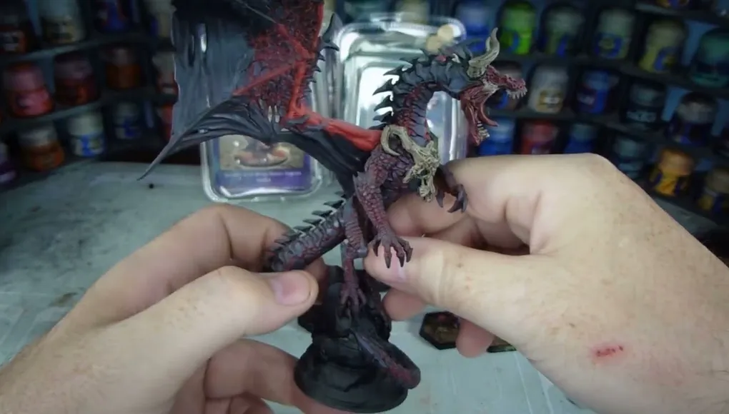 A detailed dragon miniature is being held in a hand, showcasing its intricate features, including prominent scales, fierce facial expression, and large wings with a red underside. The dragon is painted in dark hues with highlights that accentuate its muscular form and menacing appearance. In the background, various paint jars are visible, indicating a workspace for model painting.
