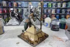Miniature figurine of a feline humanoid with green eyes, brown fur, and a brown vest, kneeling on a pyramid base. Staff with a crystal top in right hand. Nearby: two paint pots, one labeled "Citadel Colour Base Corvus Black." Background shelves hold more figurines and supplies.