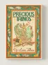 The cover of "Precious Things" features a whimsical illustration of two small dragons nestled together, surrounded by various mundane objects like buttons, a spool of thread, and dried flowers. The title "PRECIOUS THINGS" is prominently displayed at the top in a playful font, with decorative elements framing the image. The overall color palette includes warm tones of green, orange, and cream, creating a cozy and inviting aesthetic.