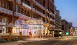 A grand hotel entrance features an ornate glass canopy with decorative lighting, flanked by brickwork and balconies of the building. The DoubleTree Metropole Hotel is prominently displayed above the entrance, while flower pots add a touch of color to the scene. The street is quiet, illuminated by soft evening lights, creating a serene atmosphere as dusk settles.
