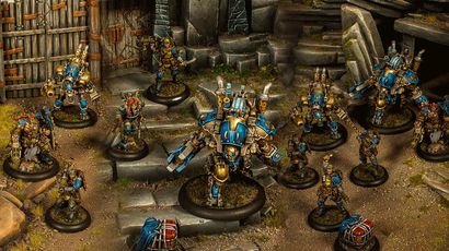 A group of Cygnar miniatures is depicted in a detailed tabletop setting, showcasing the Gravedigger Auxiliaries unit. In the foreground, a character heavy warjack named Beaker stands prominently, adorned in blue and gold armor, with various mechanical features. Surrounding it are several elite veterans, including a female commander with red hair and a rifle, an ogrun captain, and a unit of Gravedigger Commandos, all featuring a mix of blue and gold uniforms, set against a backdrop of stone steps and a weathered door.
