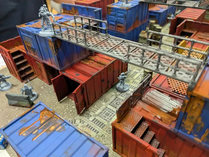 Angled overhead view of a detailed miniature tabletop terrain model. The scene depicts a multi-level industrial or sci-fi setting made of modular components. Visible elements include:

- Stacked shipping containers painted in blue, red, and weathered brown, some with open doors.
- Elevated metal walkways with railings connecting different sections.
- Small figurines, including one resembling a stormtrooper standing on a blue container and another near a control panel.
- Various industrial structures, including a blue cabinet with drawers, a wooden crate, and a metal barrel.
- A patterned floor resembling a grid or technical layout.
- No readable text is visible.