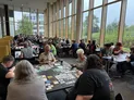 A bustling board game convention scene at the University of Northampton, featuring numerous tables filled with players engaged in various games. In the foreground, a group of four players is focused on a game spread across their table, with colorful game components and cards visible. Large windows in the background allow natural light to fill the space, highlighting the many attendees seated at tables throughout the room.