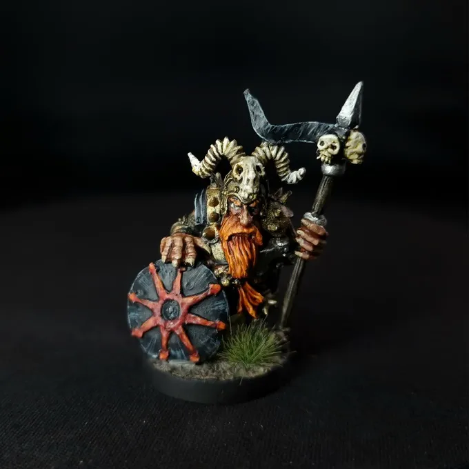 A painted miniature figure of a Chaos Dwarf warrior with a long orange beard, wearing dark armor and a helmet with large curved horns. The figure holds a round shield with a red star design and a spear topped with a skull. The base has small patches of green grass-like texture. The background is black.