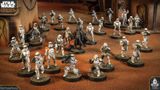 A group of Star Wars miniatures features several Stormtroopers in white armor, positioned in a desert-like environment with a backdrop of a weathered building. Among them stands a prominent figure in black armor, holding a red lightsaber, likely representing Darth Vader. The scene is detailed with various poses of the troopers, some aiming weapons, while others appear to be in action, all set on circular bases with textured surfaces.