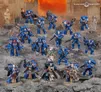 A group of Ultramarine Space Marines is displayed prominently against a rocky, war-torn backdrop. The figures are painted in vibrant blue armor, featuring various poses and weaponry, including bolters and a power sword. In the center, Chief Librarian Tigurius stands out in a flowing robe, holding a staff and flanked by several other Ultramarines, showcasing a mix of infantry types, including Bladeguard Veterans and Aggressors.