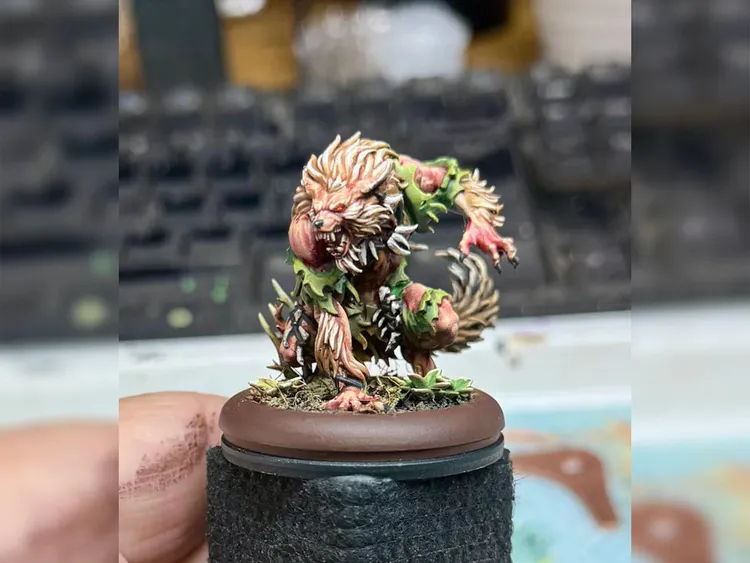 A detailed miniature of a fierce creature stands on a circular base, showcasing a blend of organic and monstrous features. The creature has a muscular build, covered in green foliage and sharp spikes, with a wild mane of hair and glowing red eyes that convey a menacing presence. Its pose suggests an aggressive stance, ready to pounce, with a background of blurred keyboard keys, indicating a workspace setting.