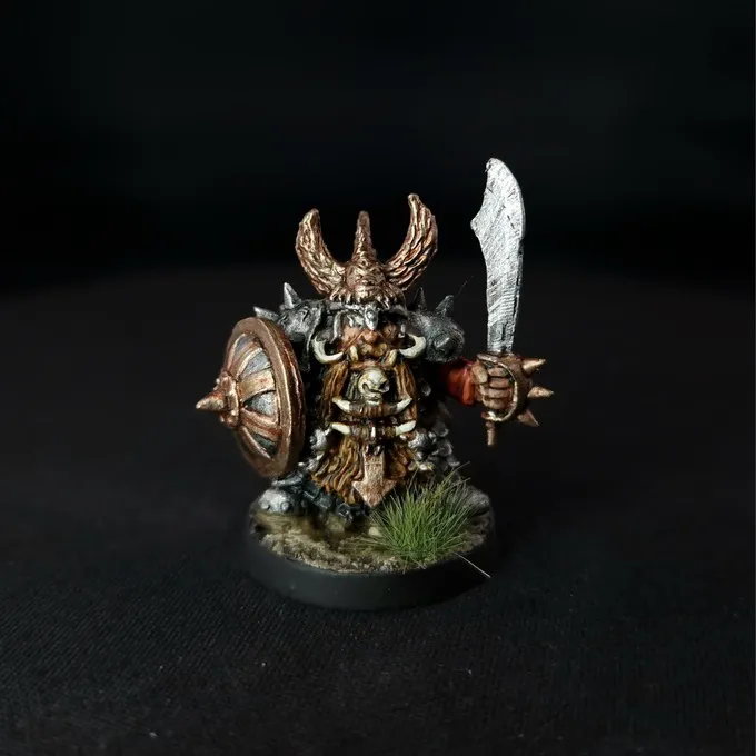 A miniature figure of a Chaos Dwarf warrior with a horned helmet, holding a large round shield with a spiked boss on the left arm and a sword in the right hand. The figure has a long braided beard, detailed armor, and is standing on a small circular base with patches of grass. The background is black.