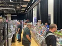 A bustling convention scene at Britcon features numerous attendees browsing a variety of game-related merchandise displayed on tables. The setup includes stacks of game boxes, rulebooks, and promotional materials, with people engaged in conversation and exploration throughout the space. The venue has a modern design, highlighted by black curtains and a polished wooden floor, creating an inviting atmosphere for historical wargaming enthusiasts.