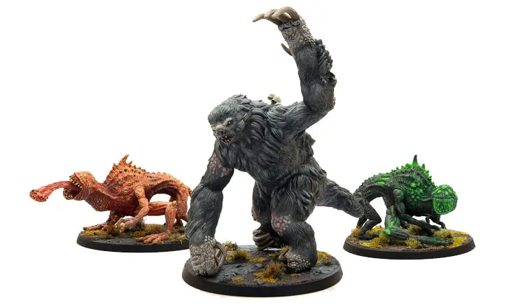 The image features three miniatures from Modiphius’ Fallout: Wasteland Warfare line. On the left is a snallygaster, a hexapod creature with a reddish-brown body, multiple eyes on its back, and a long, protruding tongue. In the center stands a large, muscular megasloth, covered in gray fur, with a menacing pose and a raised arm. To the right is another creature, possibly a variant of the snallygaster, in green, with a textured body and large eyes, all set on circular bases adorned with tufts of grass.