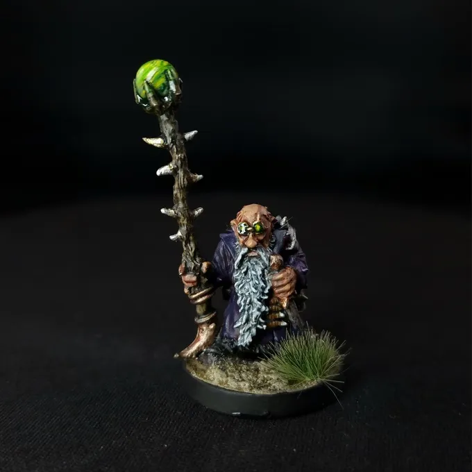 A miniature figure of a bearded dwarf wearing a dark robe, holding a staff topped with a glowing green orb. The staff is detailed with a twisted, thorn-like design. The figure has goggles over its eyes and stands on a small circular base with patches of green grass. The background is black.