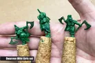 Three small, green miniatures from the "Dwarven Wild Girls" set are being held in a hand, each perched on corks. The miniatures depict stout female dwarves, each wielding an axe and showcasing distinct poses and expressions. The background is a neutral beige, allowing the details of the miniatures to stand out prominently.