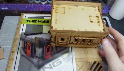 A detailed laser-cut model of a building is shown, featuring a flat roof with small square cutouts and a side with a square window and horizontal bars. The model is placed on a crafting mat with various grid patterns, and a hand is positioned near the model, indicating interaction. The overall color is a light brown, typical of wood or MDF material used in tabletop gaming terrain.