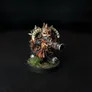 A painted miniature figure of a Chaos Dwarf with a bull-like head, curved horns, and a beard. The figure wears dark armor with orange and red fabric details, and holds a cannon-like weapon on its right shoulder. The base has green grass-like texture. The background is black.