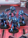 Four Sangheili warriors are depicted in striking blue armor, each wielding different weapons including a needler, plasma rifle, pulse carbine, and energy sword. They are positioned on a futuristic, metallic battlefield with a red and gray color scheme, featuring geometric structures in the background. The warriors exhibit dynamic poses, showcasing their formidable presence as mercenaries in the Halo Flashpoint universe.