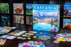 Board game box labeled "Cascadia Alpine Lakes" with mountain and lake illustration. Surrounding components include wooden trays with colorful circular tokens and round wooden discs. Other Cascadia game boxes visible in background.