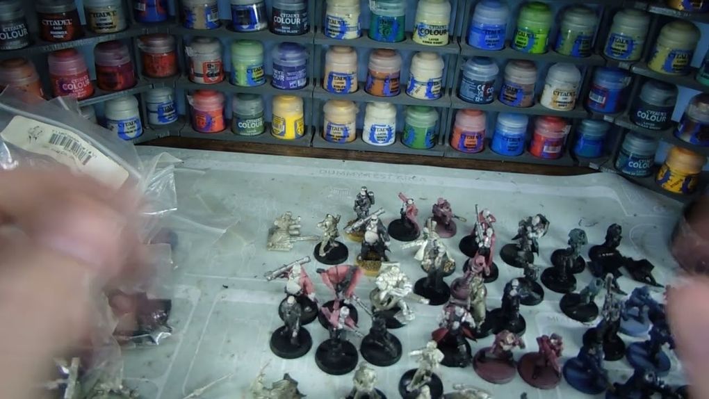 A variety of Heresy Miniatures models are arranged on a work surface, showcasing a mix of painted and unpainted figures in various poses. In the background, shelves filled with paint pots in a range of colors, including blues, greens, and reds, provide a vibrant backdrop. The models include soldiers and fantastical characters, some with detailed bases, while the surface beneath them is marked with paint splatters and tools of the hobby.