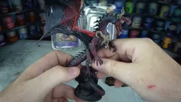 A detailed dragon miniature is being held in a hand, showcasing its intricate features, including prominent scales, fierce facial expression, and large wings with a red underside. The dragon is painted in dark hues with highlights that accentuate its muscular form and menacing appearance. In the background, various paint jars are visible, indicating a workspace for model painting.