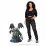 A smiling person in black hoodie, cropped top, and cargo pants stands beside a life-size statue of Cthulhu. The statue depicts a green, tentacled creature with glowing red eyes, perched on a rocky base with blue accents.