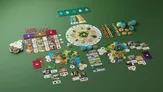 Board game components spread on green background: circular game board with radial sections, hexagonal tiles depicting landscapes, small animal and plant tokens, resource cards with icons and images of animals, plants, and terrain, and a rulebook.