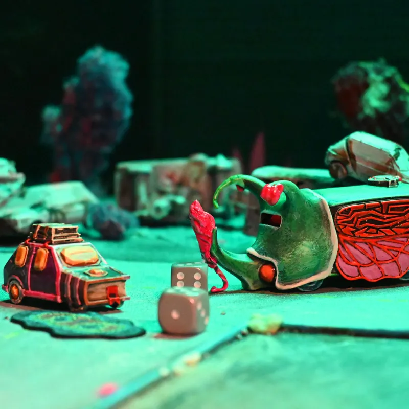 A vibrant tabletop scene features two whimsical, creatively designed vehicles: one resembling a small car with exaggerated features and a roof rack, and the other a larger, fantastical machine with a green, elongated body and intricate patterns. In the foreground, three dice are scattered on the surface, alongside a small, red object that appears to be a creature or character interacting with the larger vehicle. The background is filled with blurred, colorful debris, enhancing the chaotic atmosphere of the racing game setting.