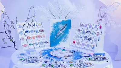 Boardgame components on a white table: Arctic wildlife cards showing fish, seals, whales, and birds; a circular game board with a blue Arctic map; hexagonal tiles with animal icons; and small circular tokens in blue, green, and purple. Central artwork depicts a blue whale.
