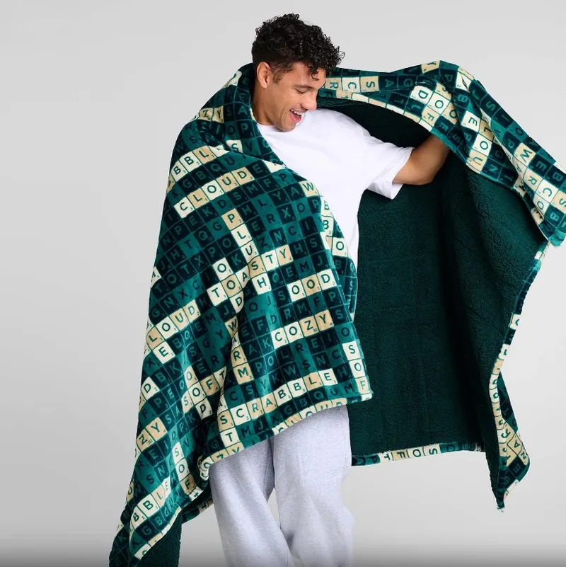Man in white shirt holding open a green Scrabble-patterned blanket.