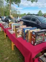 Outdoor car boot sale with red tablecloths displaying board games, DVDs, and books. People browsing and parked cars in background.