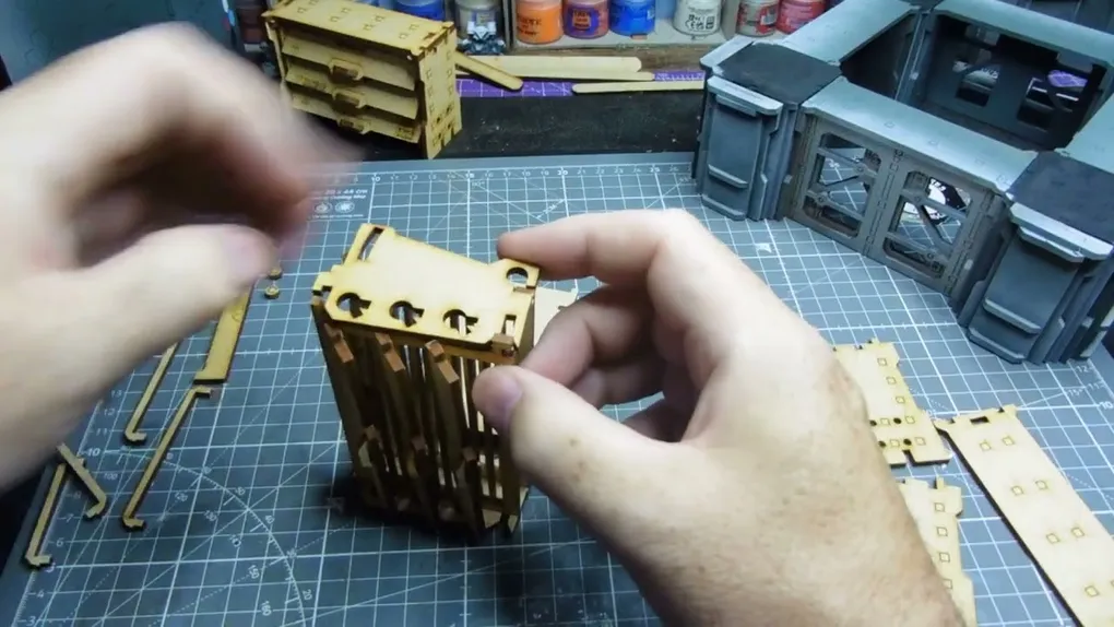 Hands assembling a wooden model on a cutting mat with tools and paint jars in the background.