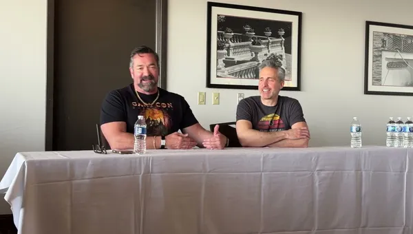Two men are seated at a table during a panel discussion. On the left, a man with a beard, wearing a black shirt featuring a colorful graphic and a necklace, gestures with his hands while speaking. On the right, a man with short gray hair, dressed in a gray t-shirt with a retro design, smiles and listens attentively. Behind them, a framed black-and-white photograph of a decorative railing is visible on the wall. A bottle of water and a small device are placed on the table in front of them.