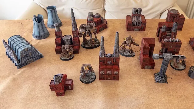 A collection of Epic scale terrain pieces suitable for games like Adeptus Titanicus and Battletech is displayed on a tan surface. The terrain includes various structures, such as a large, blue-gray building with pipes, several red industrial buildings, and tall smokestacks. Among the terrain, two detailed miniatures resembling mechs are positioned, adding to the scene's dynamic and futuristic atmosphere.