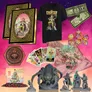 Merchant with ornate golden frame, black T-shirt with "The Painted Wastelands" and illustrated figure, playing cards featuring fantasy characters, two banknotes with fantasy designs, small figurines of humanoid and monster characters, hexagonal tile with circular emblem, and a large green stone