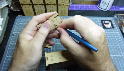 Two hands holding a partially assembled laser-cut MDF piece for a miniature building kit. The left hand supports the main structure, which is a yellowish-beige MDF panel with square cutouts and small circular holes. The right hand uses a blue-handled metal tool, likely tweezers or a small pick, to position or secure a small metal disc, possibly a magnet, into one of the circular holes. In the background, a clear plastic cutting mat with a printed grid is visible, along with several other laser-cut MDF sheets still in their frames, some with engraved or printed text and symbols. One of the MDF sheets in the background has a purple grid pattern. No readable text is visible on the main components being handled.